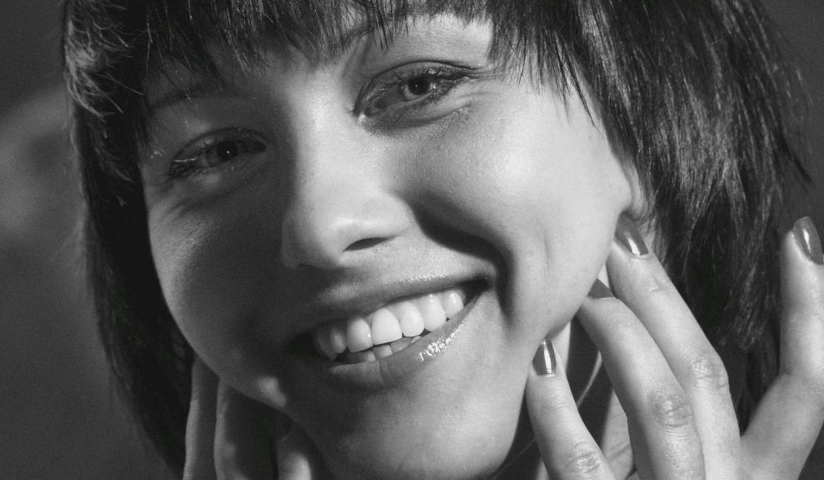 Black-and-white close-up beauty portrait of a woman with a bob haircut and bangs, smiling broadly with her hands framing her face and fingertips resting near her cheek, showcasing glowing, radiant skin.