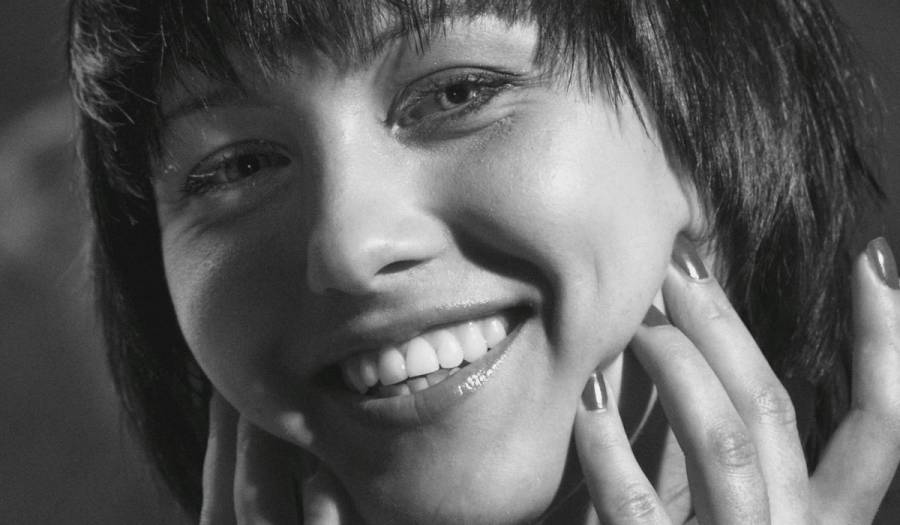 Black-and-white close-up beauty portrait of a woman with a bob haircut and bangs, smiling broadly with her hands framing her face and fingertips resting near her cheek, showcasing glowing, radiant skin.