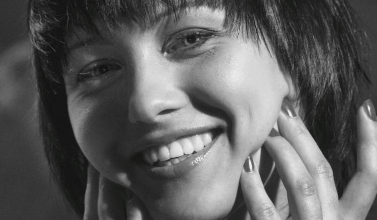 Black-and-white close-up beauty portrait of a woman with a bob haircut and bangs, smiling broadly with her hands framing her face and fingertips resting near her cheek, showcasing glowing, radiant skin.