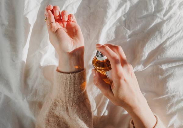 A person spritzes amber-colored perfume onto their wrist against a backdrop of soft white linen.
