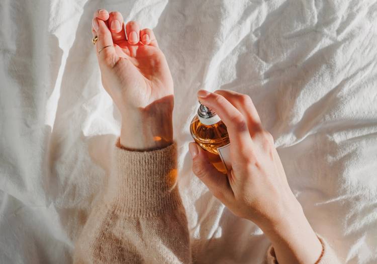 A person spritzes amber-colored perfume onto their wrist against a backdrop of soft white linen.