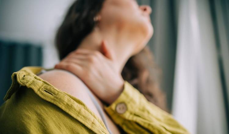 Woman in a mustard-yellow shirt tilts her head back and presses a hand to her neck, as if soothing itchy, irritated skin