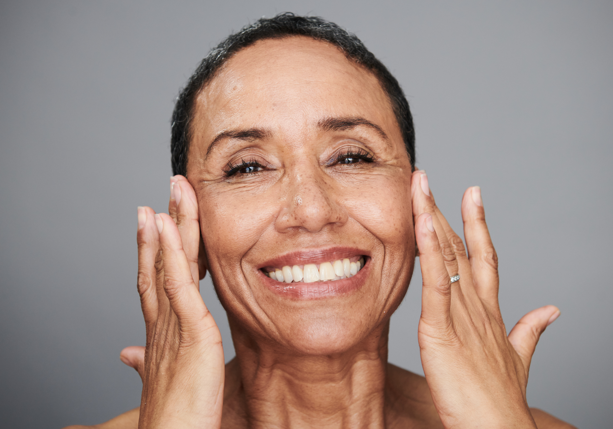 A smiling woman with mature skin touches her cheeks with both hands against a gray background