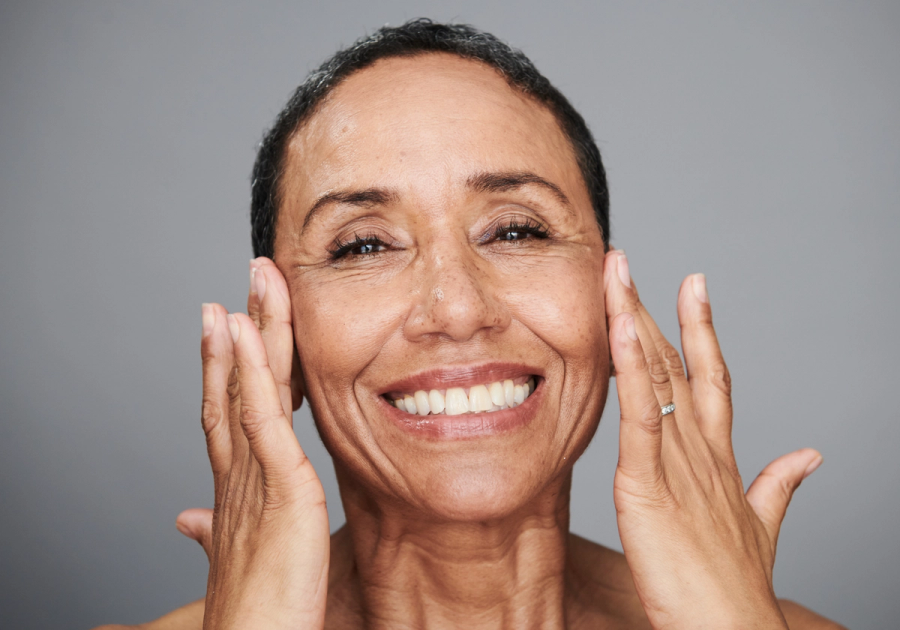A smiling woman with mature skin touches her cheeks with both hands against a gray background