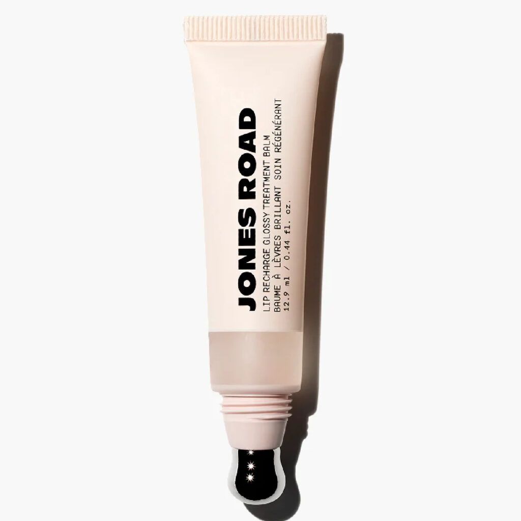 Jones Road Lip Recharge Glossy Treatment Balm in a beige tube with a sheer neutral formula visible through a translucent window and a metal applicator tip against a light gray background.