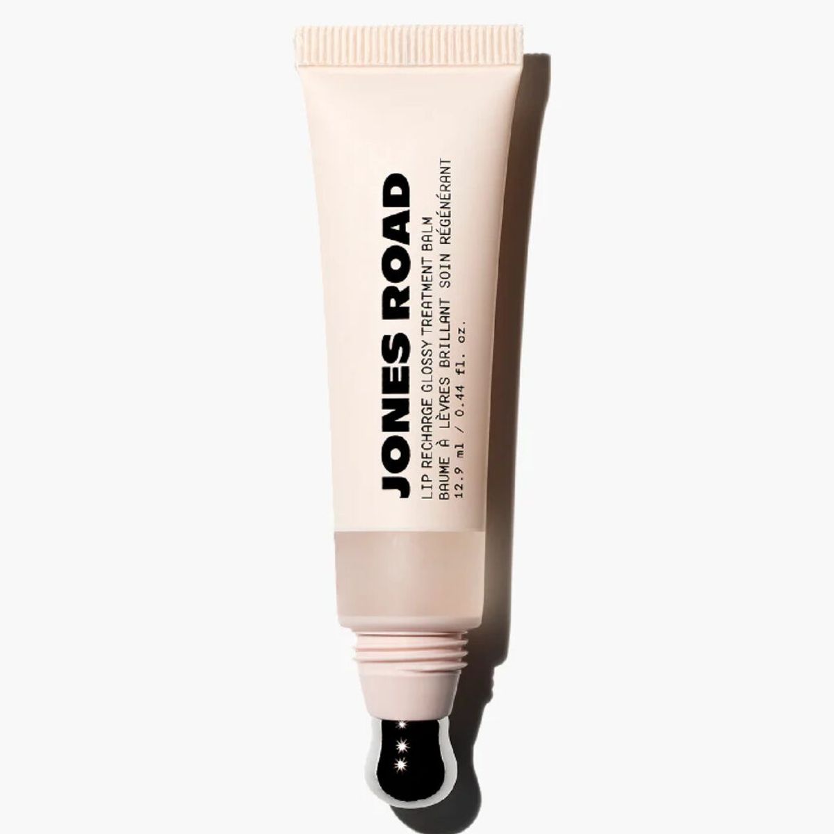 Jones Road Lip Recharge Glossy Treatment Balm in a beige tube with a sheer neutral formula visible through a translucent window and a metal applicator tip against a light gray background.