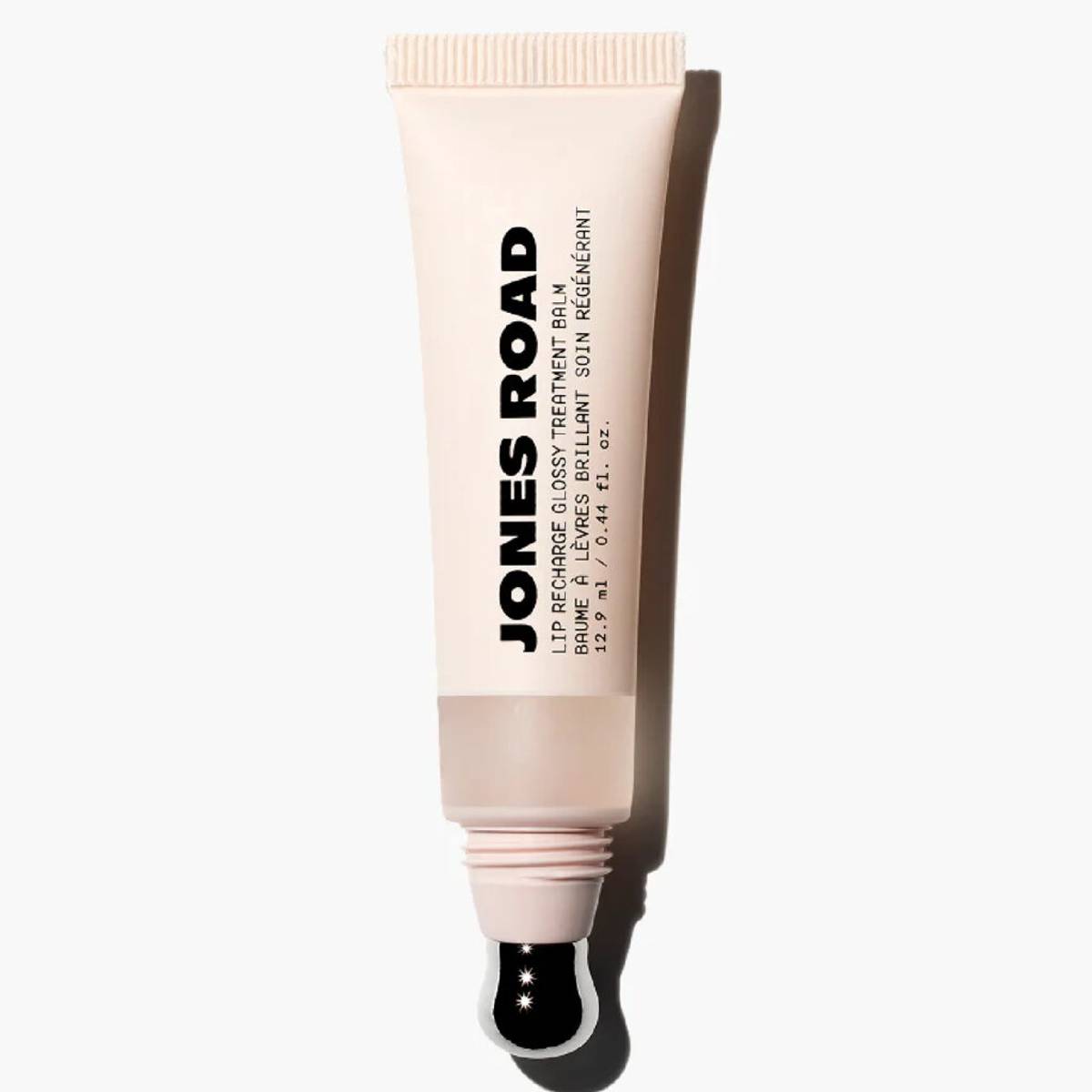 Jones Road Lip Recharge Glossy Treatment Balm in a beige tube with a sheer neutral formula visible through a translucent window and a metal applicator tip against a light gray background.