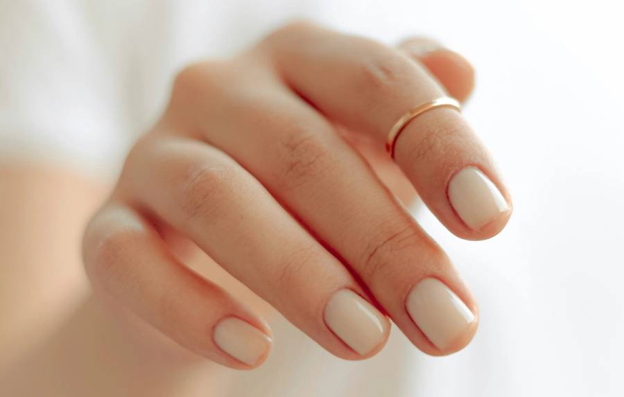 Close-up of a hand with short, glossy white nails and a thin gold ring against a softly blurred background.