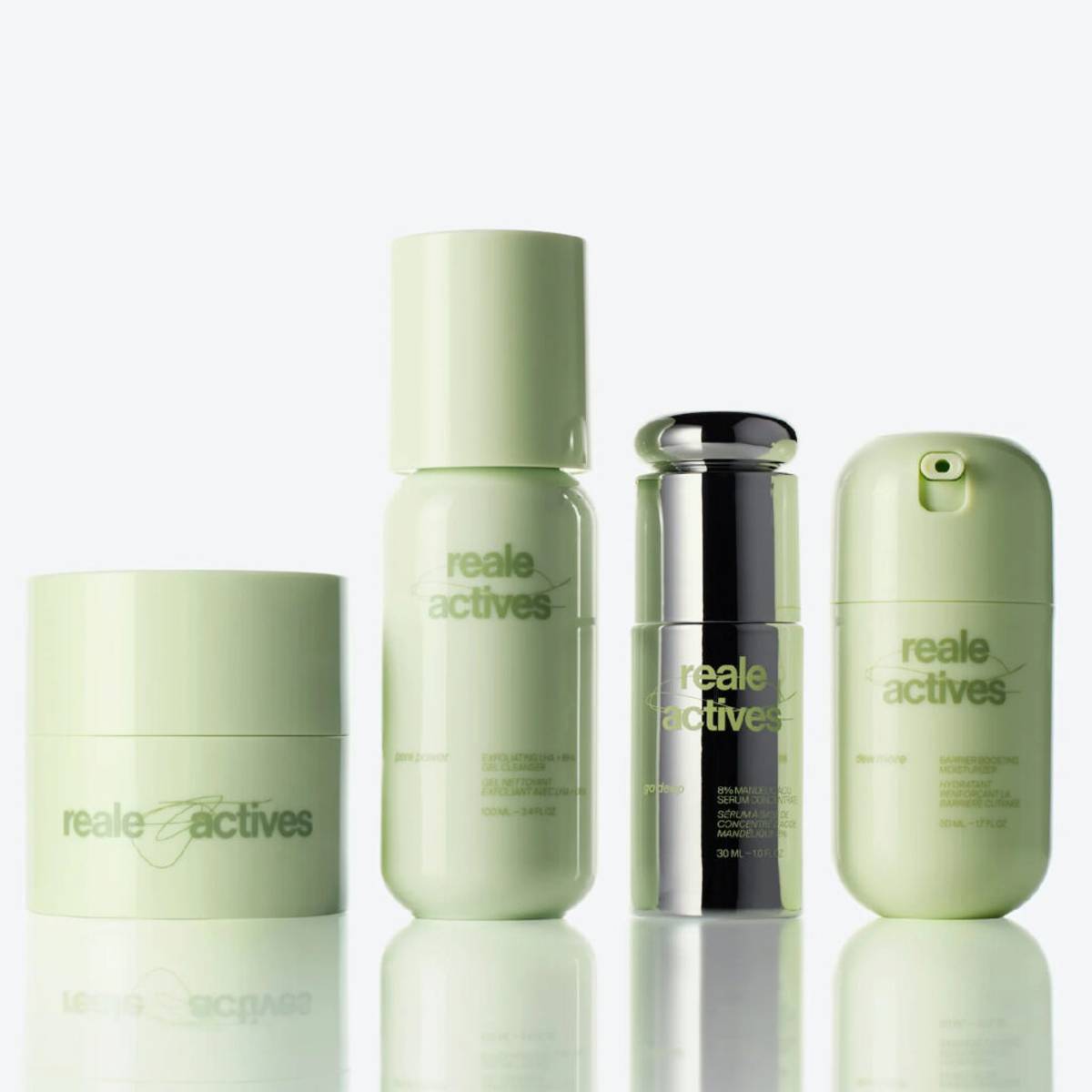 Reale Actives skin-care collection featuring four products—a cleansing balm, exfoliating gel cleanser, mandelic acid serum and barrier-boosting moisturizer—in pale green and chrome packaging, displayed on a glossy surface against a white background.