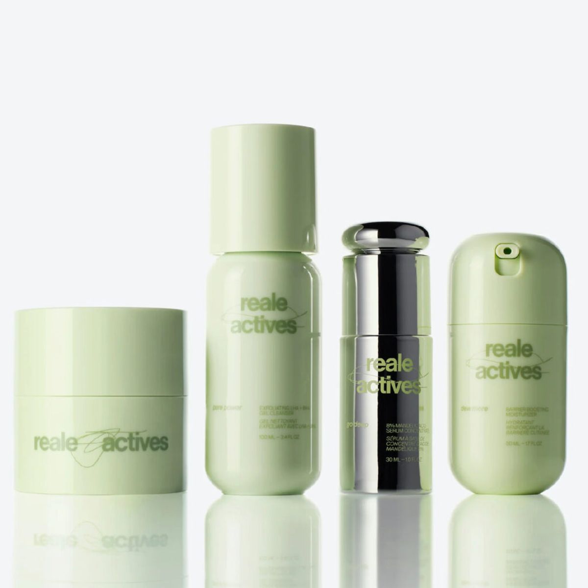 Reale Actives skin-care collection featuring four products—a cleansing balm, exfoliating gel cleanser, mandelic acid serum and barrier-boosting moisturizer—in pale green and chrome packaging, displayed on a glossy surface against a white background.