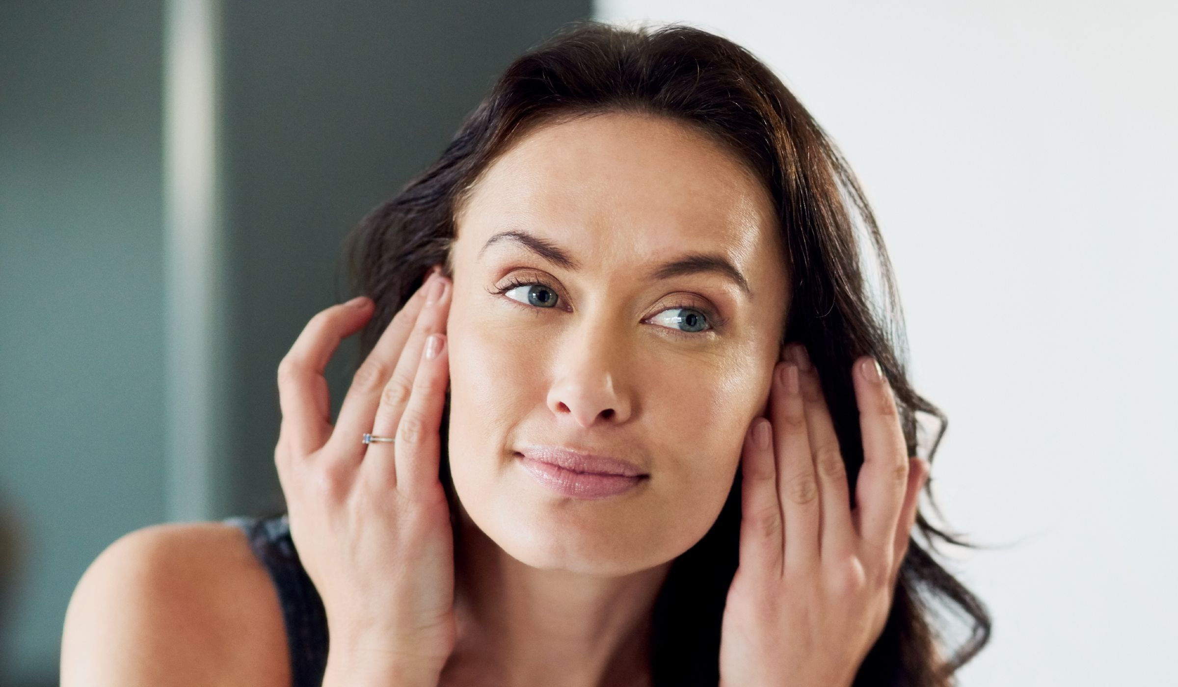 Woman examining her face in a mirror with both hands near her temples