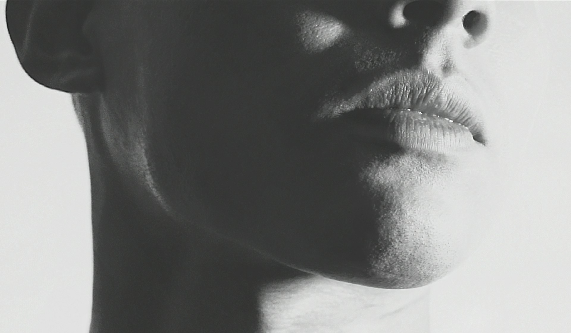 Black-and-white close-up profile shot of a person's lower face, focused on the lips, chin, and jawline, with dramatic high-contrast lighting