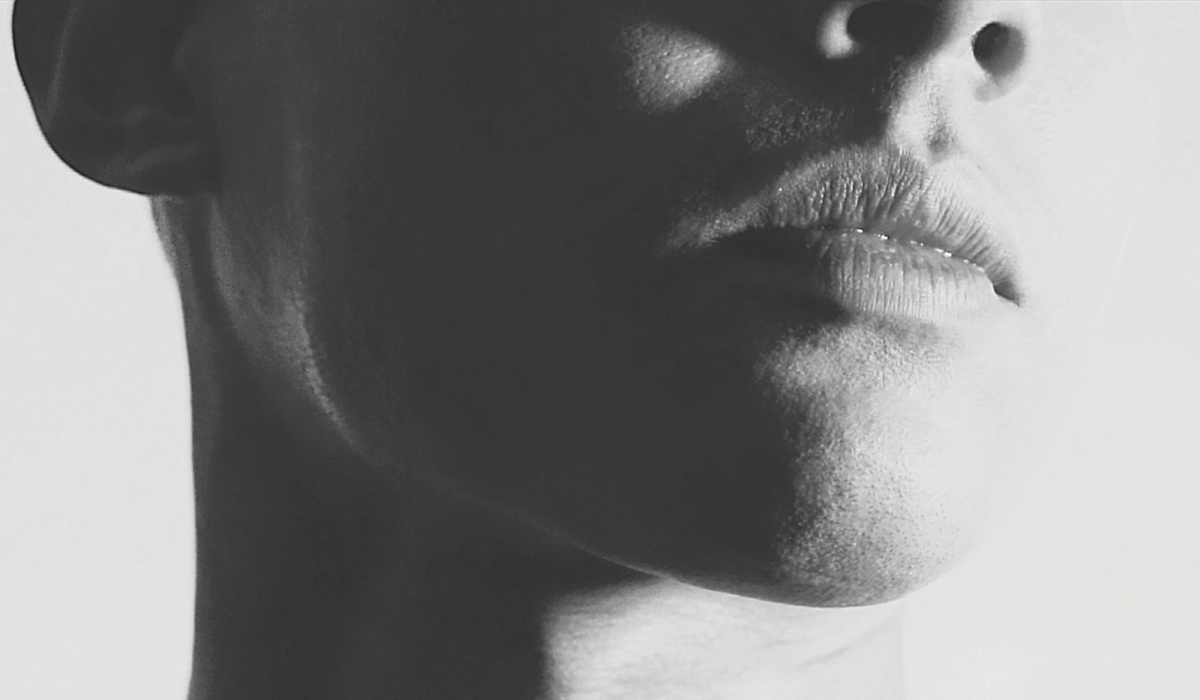 Black-and-white close-up profile shot of a person's lower face, focused on the lips, chin, and jawline, with dramatic high-contrast lighting
