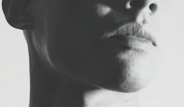 Black-and-white close-up profile shot of a person's lower face, focused on the lips, chin, and jawline, with dramatic high-contrast lighting