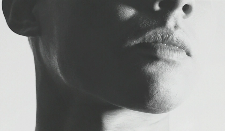 Black-and-white close-up profile shot of a person's lower face, focused on the lips, chin, and jawline, with dramatic high-contrast lighting