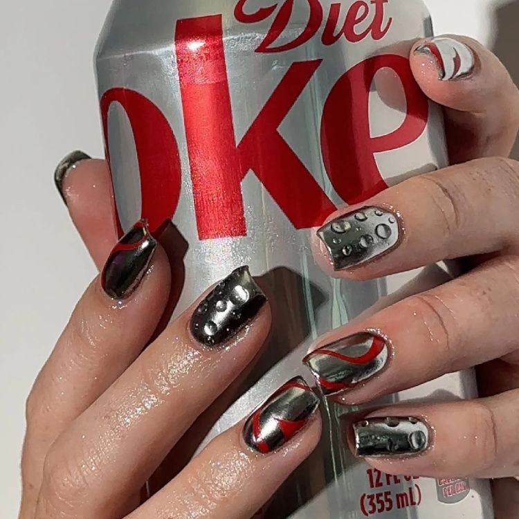 Diet Coke–inspired nail art featuring chrome silver nails with red swirl accents, soda droplet details and glossy metallic finishes, shown holding a Diet Coke can