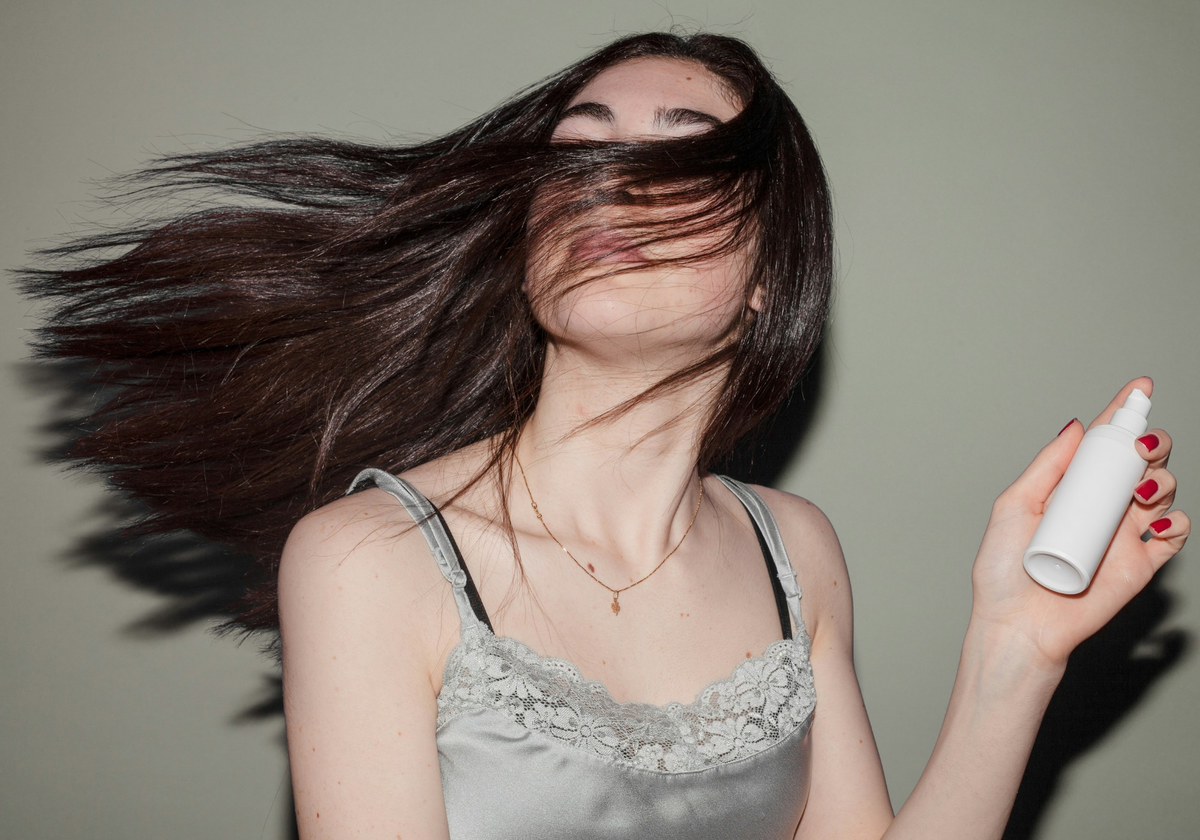 Woman with long brown hair flipping across her face while holding a white dry shampoo bottle.