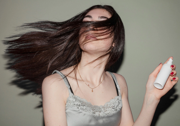 Woman with long brown hair flipping across her face while holding a white dry shampoo bottle.