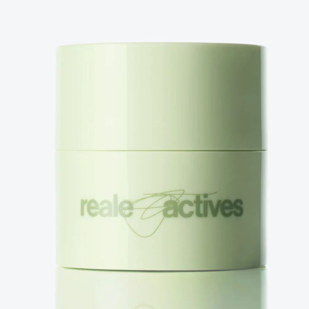 Reale Actives cleansing balm in a pale green cylindrical jar with a minimalist design, reflected on a glossy surface against a clean white background.