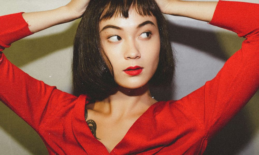 Alt text: Woman with a sleek dark bob and short micro bangs poses with arms raised, wearing a red top and bold red lipstick against a neutral background