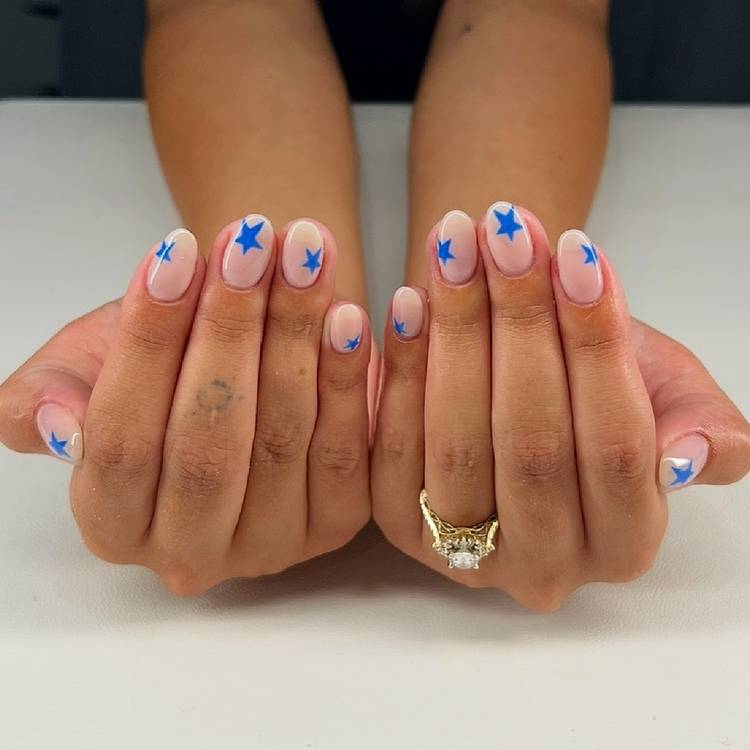 Close-up of two hands resting on a white surface with sheer nude oval nails featuring bold cobalt blue star nail art on every finger, with a diamond ring visib…Close-up of two hands resting on a white surface with sheer nude oval nails featuring bold cobalt blue star nail art on every finger, with a diamond ring visible on one hand