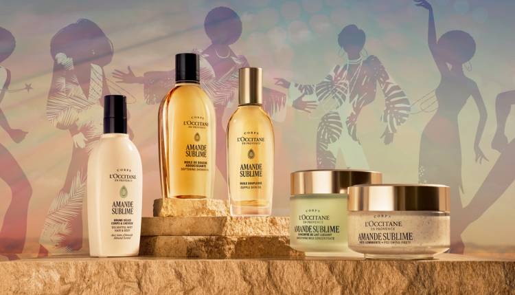 L’Occitane en Provence Almond collection products displayed on a stone surface against a warm, sunset-colored backdrop with silhouetted dancing figures.