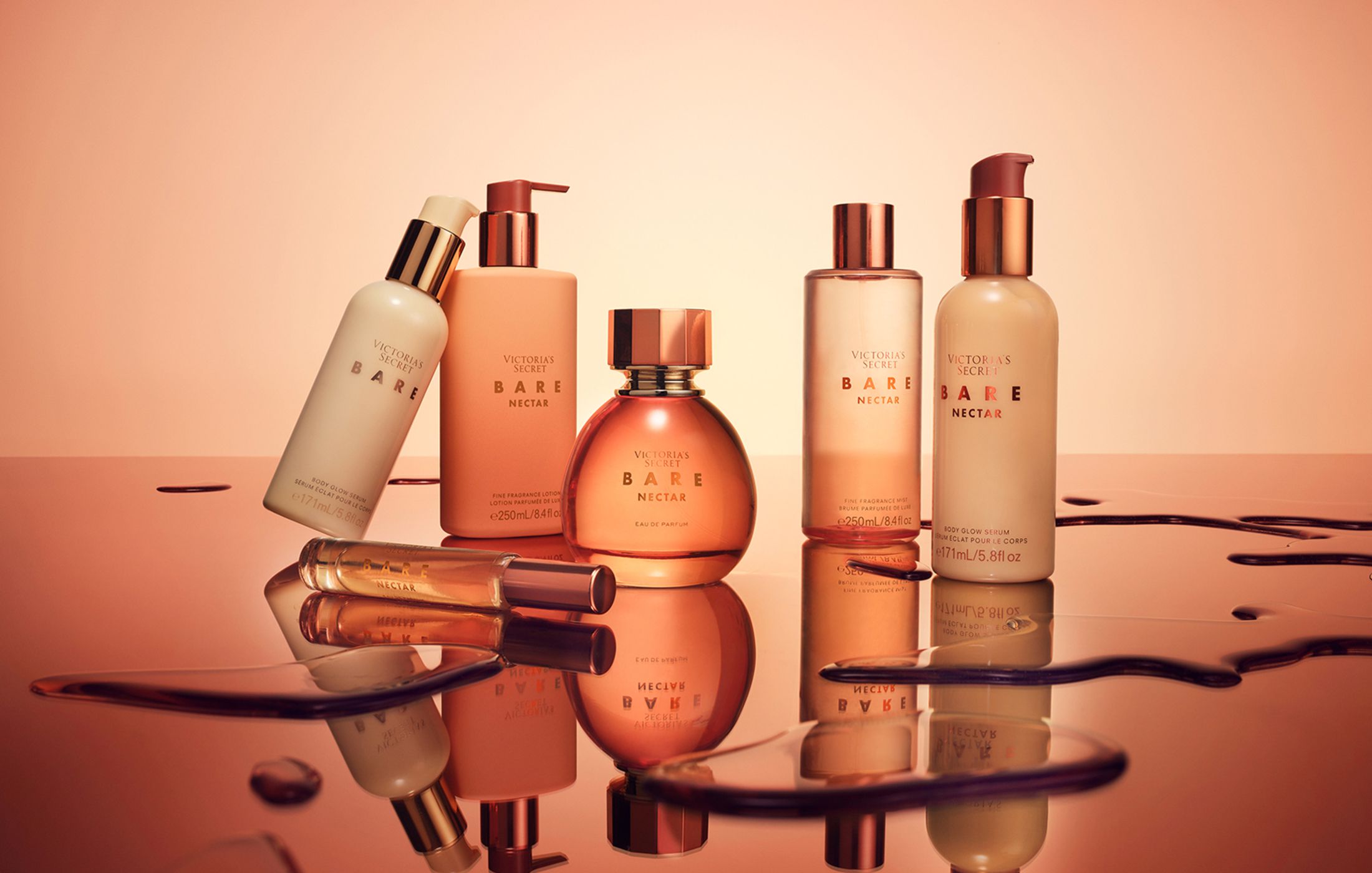 Victoria's Secret Bare Nectar collection, including body glow serum, fragrance lotion, eau de parfum and fragrance mist, arranged on a reflective surface with a warm peach-toned background.