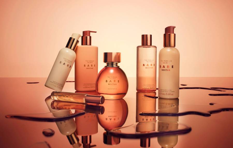 Victoria's Secret Bare Nectar collection, including body glow serum, fragrance lotion, eau de parfum and fragrance mist, arranged on a reflective surface with a warm peach-toned background.