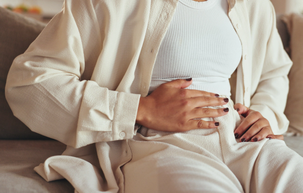 Woman in cream loungewear sitting on a couch with her hand resting on her stomach, gesturing toward menstrual cramps