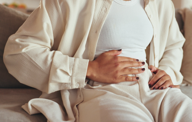 Woman in cream loungewear sitting on a couch with her hand resting on her stomach, gesturing toward menstrual cramps
