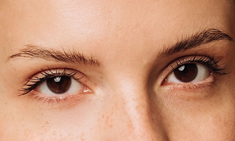 extreme close up of female brown eyes with clear skin eyebrows eyelashes and freckles looking at camera
