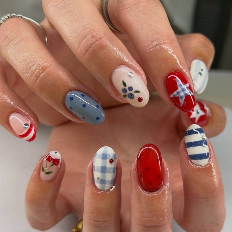 Close-up of two hands with an intricate mixed pattern manicure featuring blue gingham, red and white stripes, blueberry nail art, red floral accents, a gingham star on red, blue polka dots and navy stripe nails with rhinestone details on almond-shaped nails