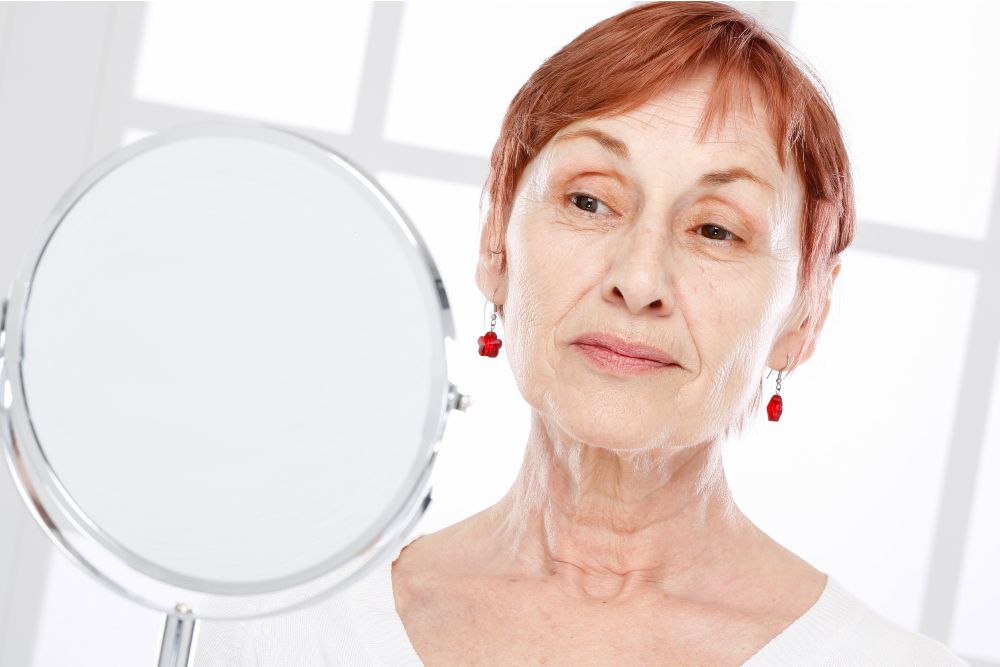 Older woman looking into a mirror, examining facial contours and skin as an illustration for a story on how facelift results settle and evolve over time.