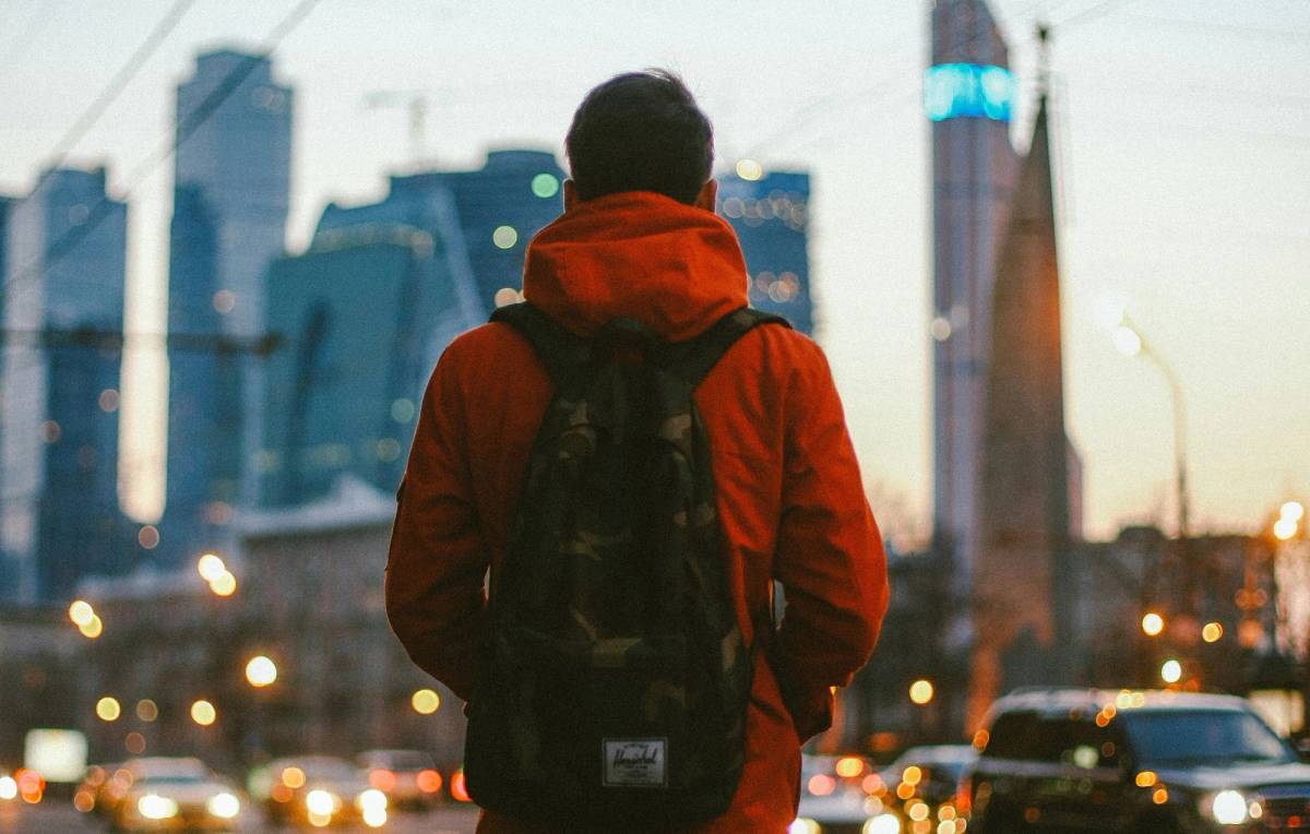 A man in an orange hooded jacket, seen from behind with a backpack, faces a blurred city skyline of high-rise buildings at dusk with traffic and street lights below.