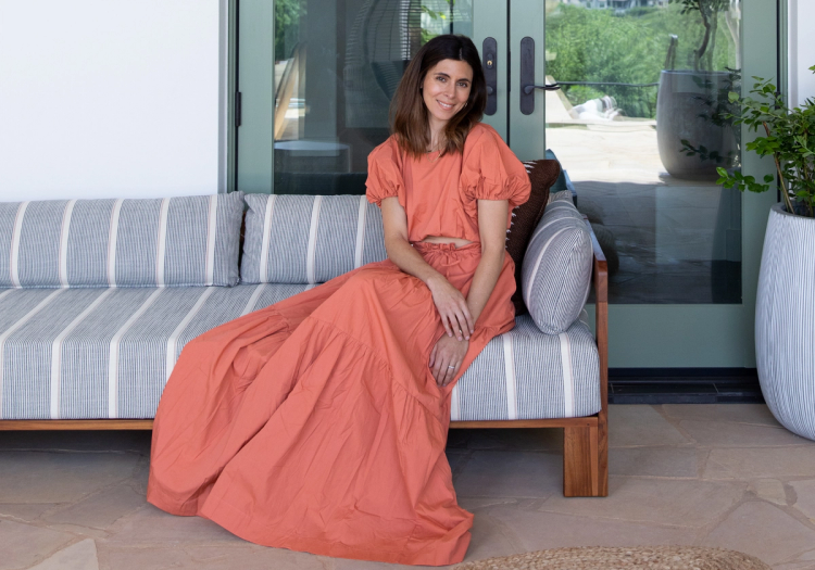 Jamie-Lynn Sigler sitting on a striped outdoor sofa in a coral puff-sleeve top and matching skirt, smiling in front of glass patio doors with greenery in the background.