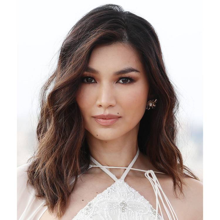 Close view of Gemma Chan who's wearing a white halter dress and wavy medium-length brown hair