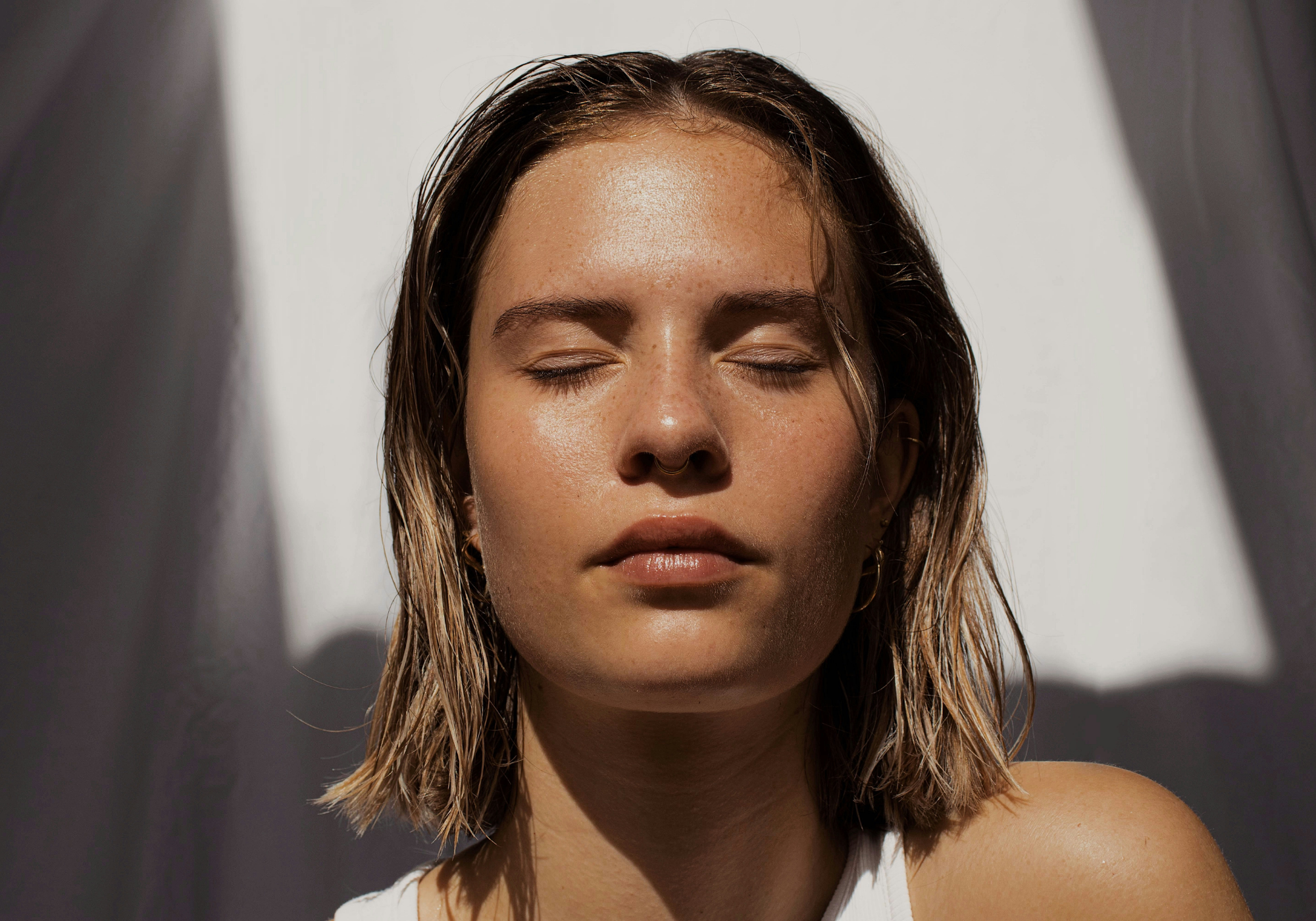 Close-up portrait of a woman with wet hair and closed eyes, sunlight highlighting bare, dewy skin.