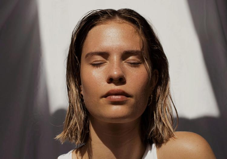 Close-up portrait of a woman with wet hair and closed eyes, sunlight highlighting bare, dewy skin.