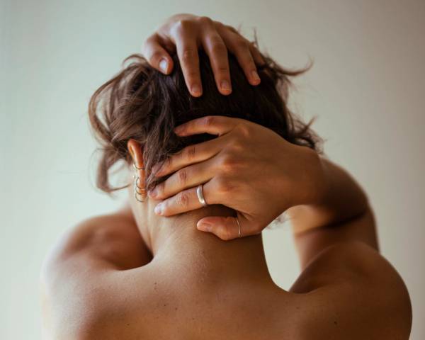 A woman seen from behind with bare shoulders, hands in her tousled brown hair, against a soft pale background.