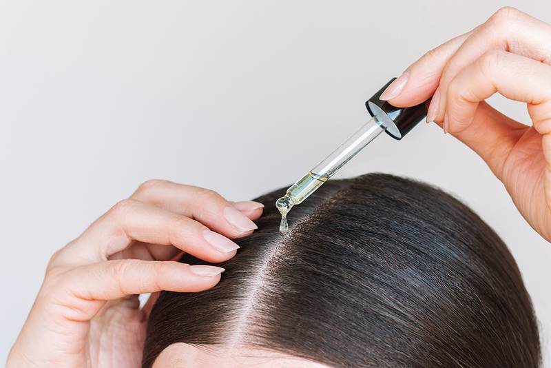 Close-up young brunette woman applying a drop of oil from pipette to her scalp. Hair loss. Strengthening and growth of hair. Hair care.