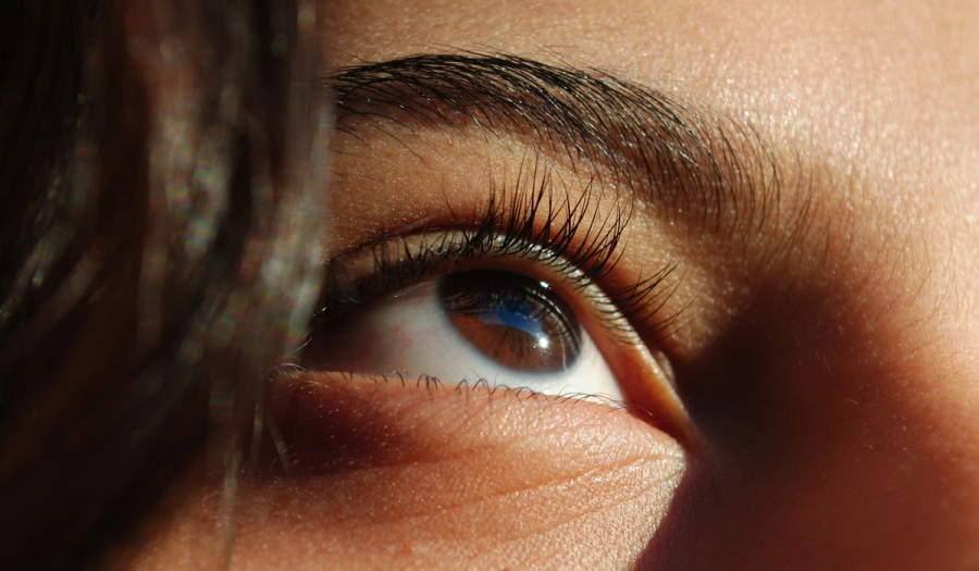Extreme close-up of a woman's brown eye and eyebrow, with defined lashes and light catching the skin's texture, gazing upward.