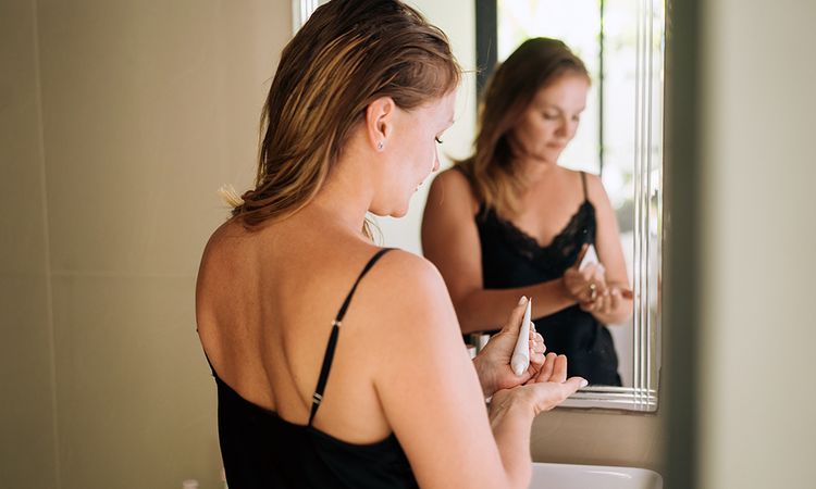 A beautiful middle-aged woman in a nightgown applies cosmetics in the bathroom near the mirror. Daily morning routine, facial skin care, home treatment. A room in a luxury hotel, business trip or vacation in a high class apartment. Relaxation, mindfulness and zen-like