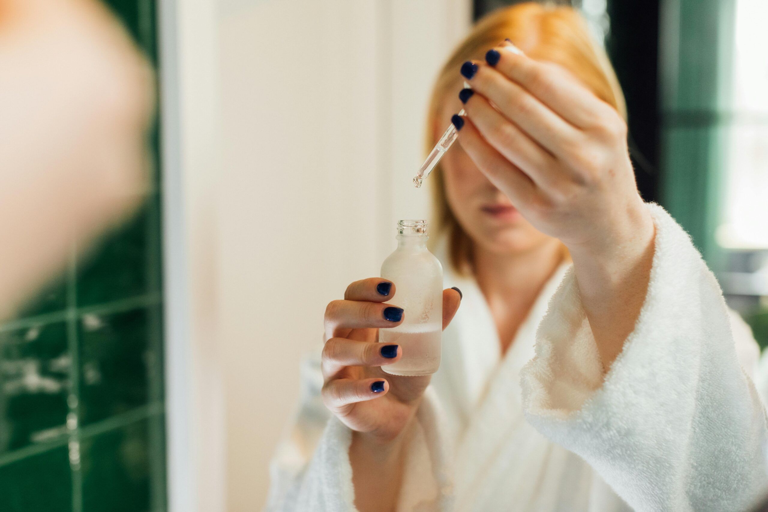 Woman using a dropper to apply serum as part of her skincare routine in a bathroom setting.