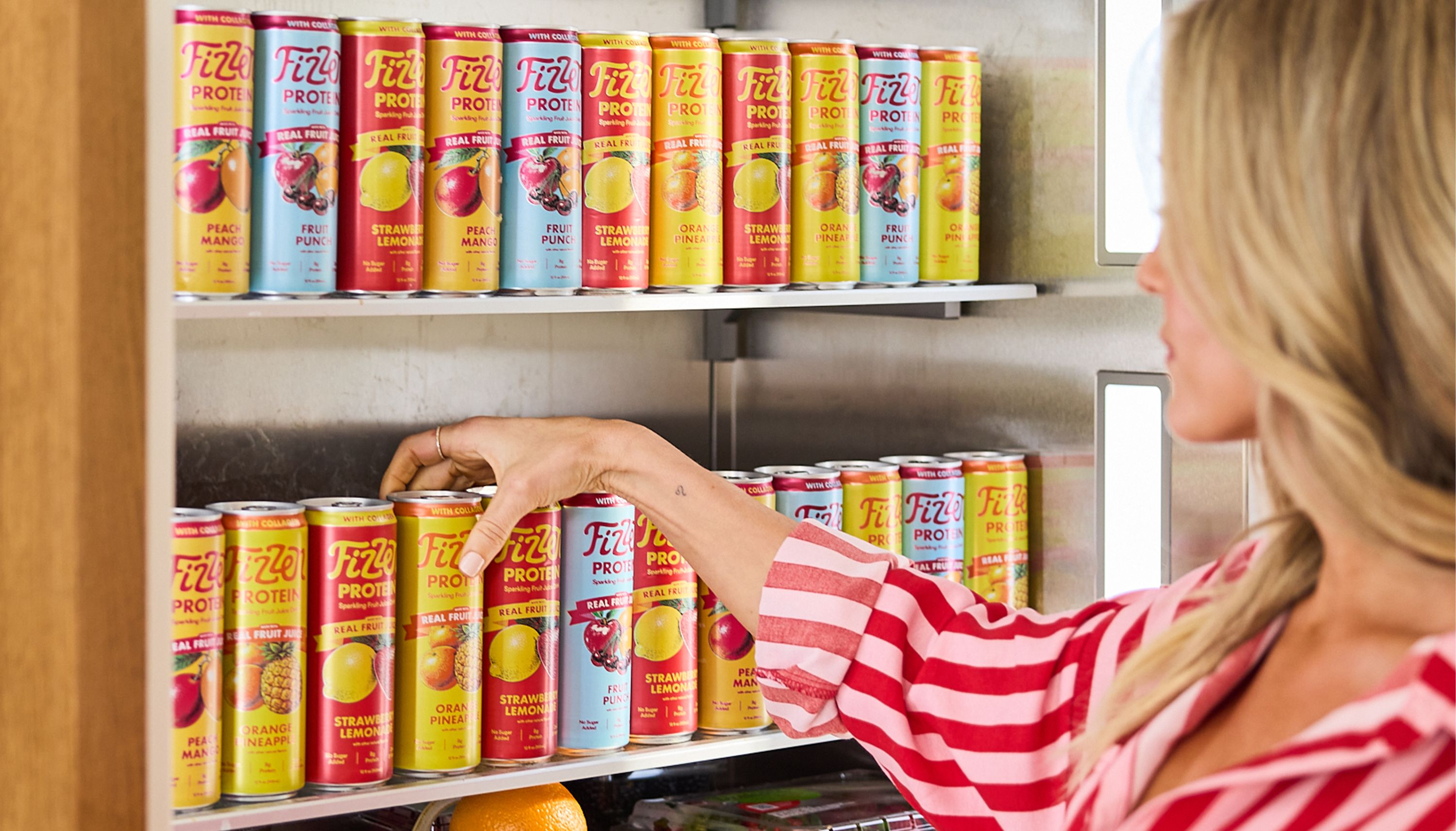 A woman in a pink striped shirt reaches for a can of Fizzen Protein sparkling drink from a refrigerator shelf stocked with all four flavors: Peach Mango, Fruit Punch, Strawberry Lemonade and Orange Pineapple.