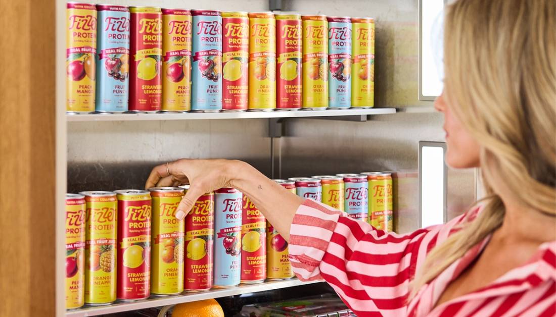 A woman in a pink striped shirt reaches for a can of Fizzen Protein sparkling drink from a refrigerator shelf stocked with all four flavors: Peach Mango, Fruit Punch, Strawberry Lemonade and Orange Pineapple.