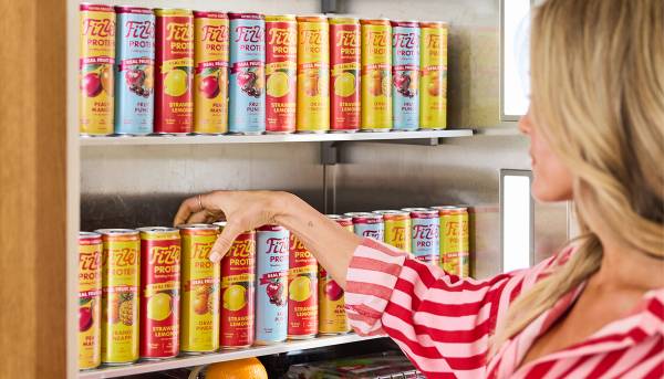 A woman in a pink striped shirt reaches for a can of Fizzen Protein sparkling drink from a refrigerator shelf stocked with all four flavors: Peach Mango, Fruit Punch, Strawberry Lemonade and Orange Pineapple.