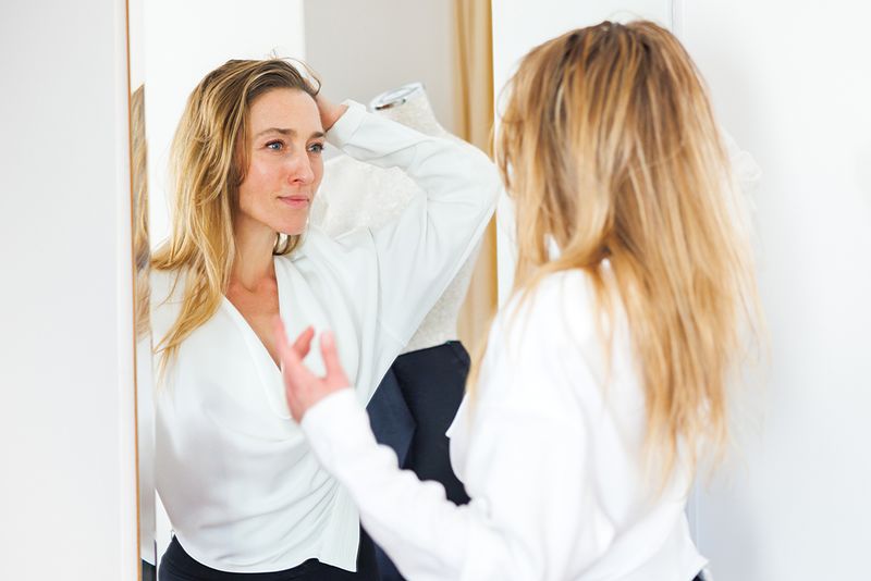 Woman wearing a newly designed white shirt, looking at her own reflection in the mirror with hand in hair, feeling proud of her work