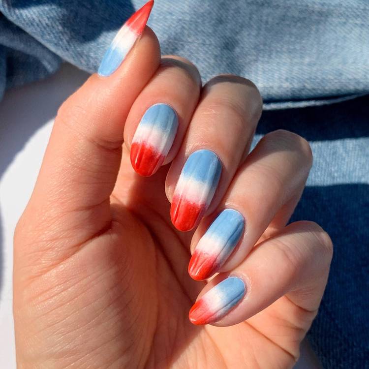 Close-up of a hand with almond-shaped nails featuring a red, white and blue ombre gradient design, photographed in sunlight against a denim background