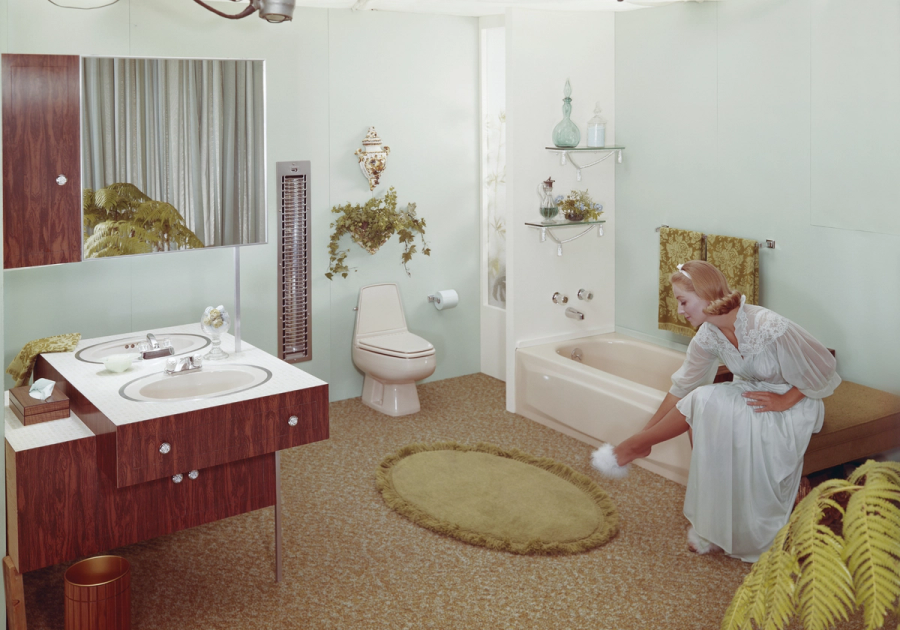 A woman in a white robe sits in a retro bathroom, reaching toward a fluffy white slipper beside a bathtub, vanity and green shag rug.