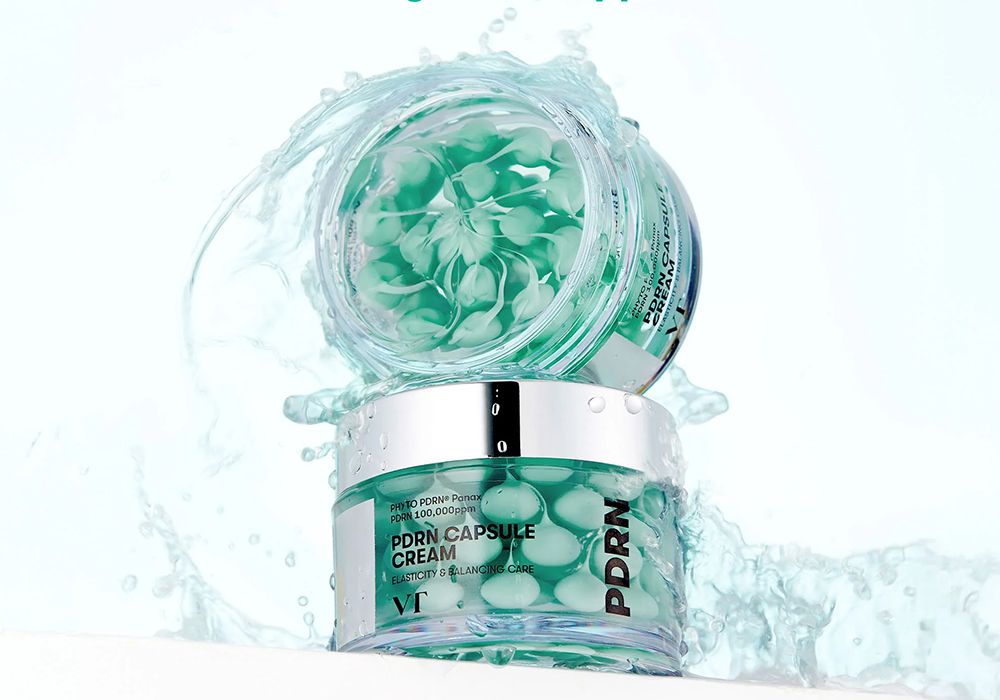 An open jar of VT Cosmetics PDRN Capsule Cream showing green gel capsules inside, surrounded by water splashes against a light blue background.